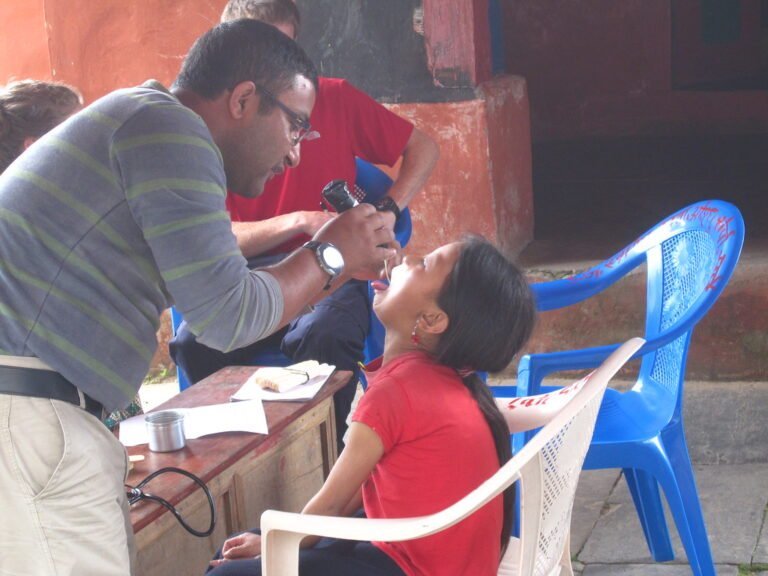 A man in a striped sweater is examining a young girl's eyes while she sits in a plastic chair, tilting her head back. They appear to be outdoors and part of a vision screening event.