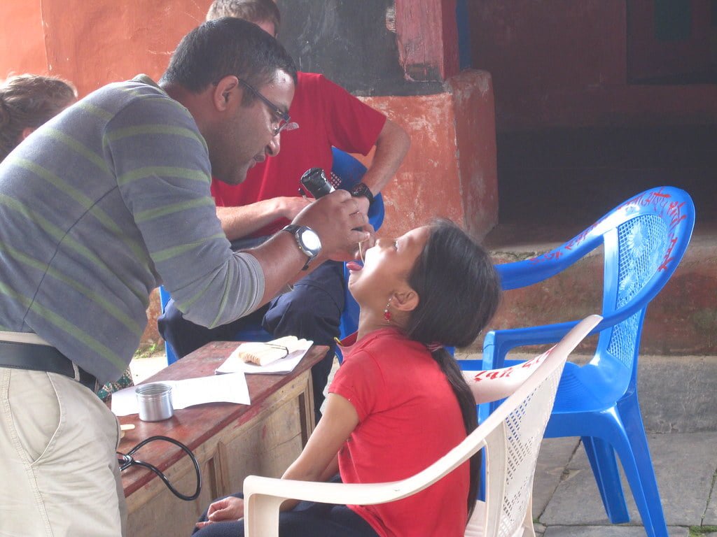 A man in a striped sweater is examining a young girl's eyes while she sits in a plastic chair, tilting her head back. They appear to be outdoors and part of a vision screening event.