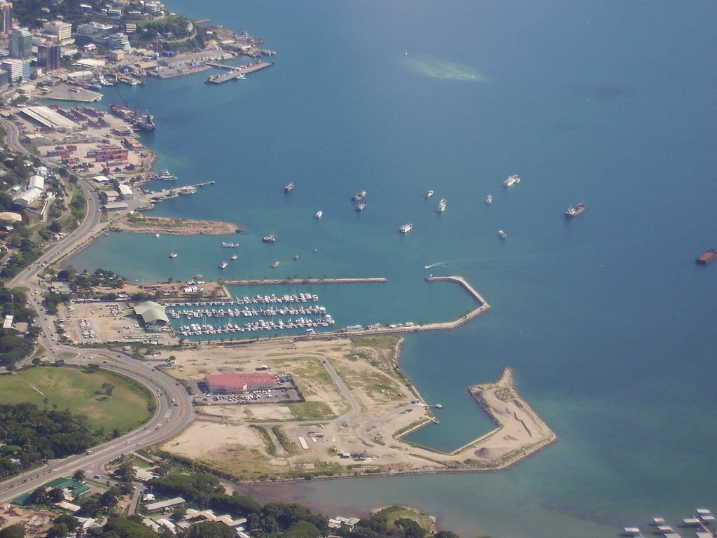 Aerial view of a coastal area with a marina, various ships at anchor, and urban development near the shoreline.