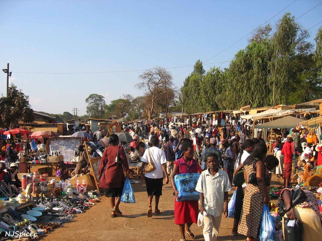 A bustling open-air market with numerous vendors and shoppers, offering a variety of goods under a clear sky.