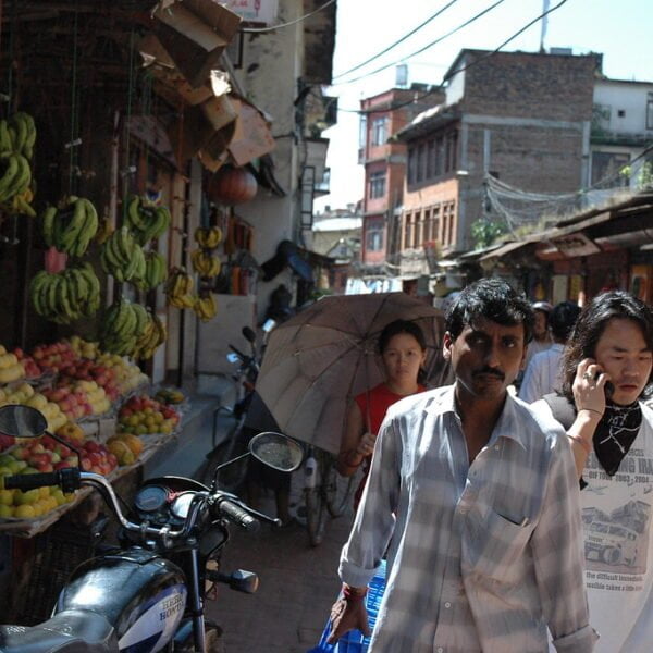 A bustling street scene with people walking, a fruit market on the left with bananas and other fruits on display, motorcycles parked on the side, and traditional buildings with various signs and electrical wires overhead.