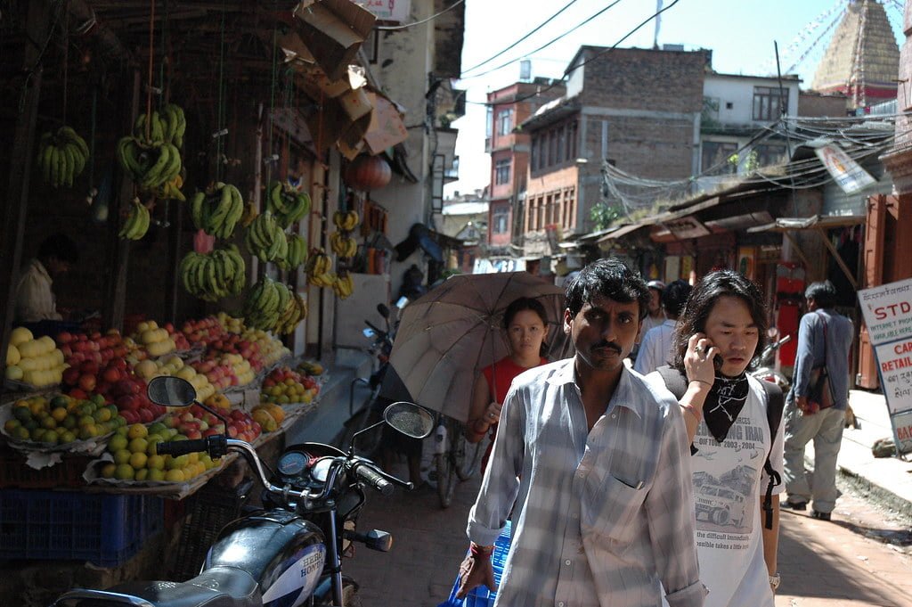 A bustling street scene with people walking, a fruit market on the left with bananas and other fruits on display, motorcycles parked on the side, and traditional buildings with various signs and electrical wires overhead.