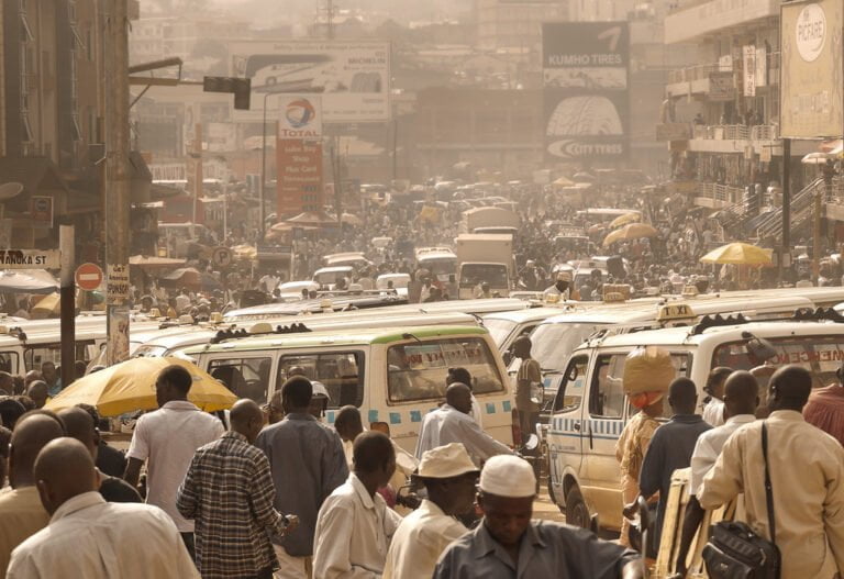 A bustling street scene with heavy pedestrian and vehicular traffic, including numerous white vans with colorful stripes and several people carrying yellow umbrellas. Signage for businesses and tires appear in the background amidst a hazy, sunlit atmosphere.