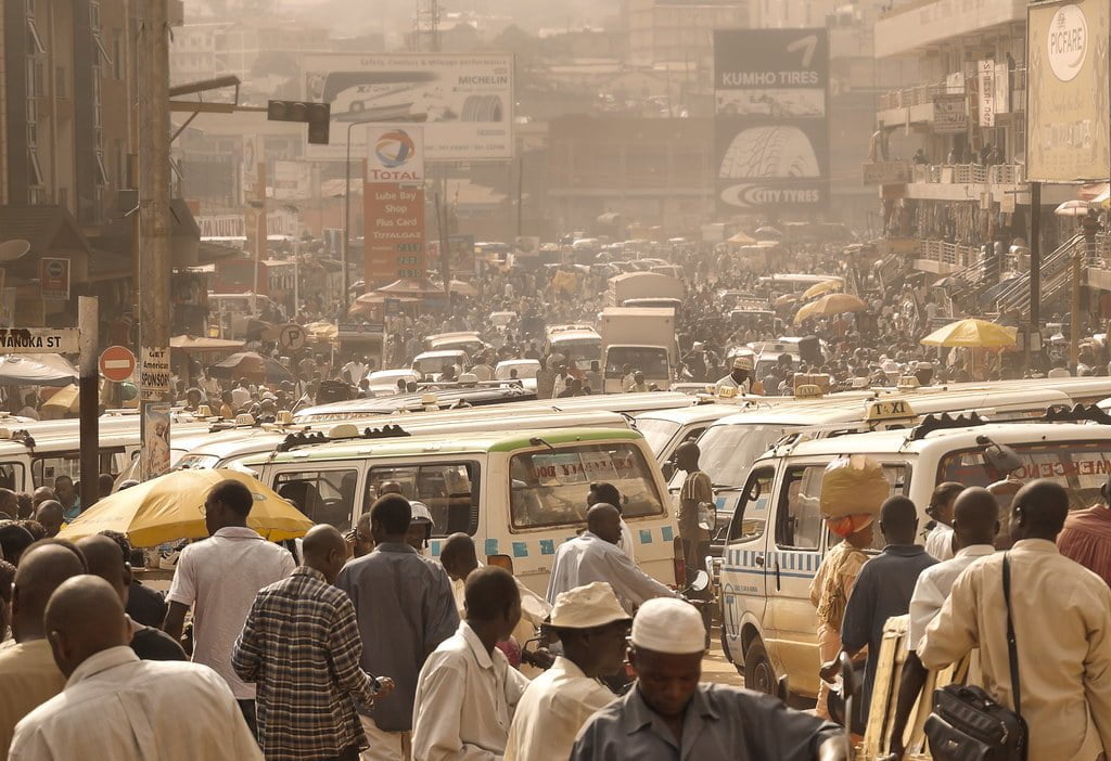 A bustling street scene with heavy pedestrian and vehicular traffic, including numerous white vans with colorful stripes and several people carrying yellow umbrellas. Signage for businesses and tires appear in the background amidst a hazy, sunlit atmosphere.