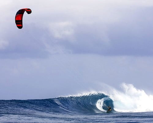 A kite surfer riding a large wave with a red and black kite aloft in a cloudy sky.