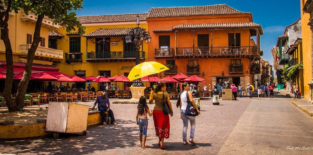 A vibrant street scene with pedestrians, colorful colonial buildings, outdoor seating under umbrellas, and a clear blue sky.