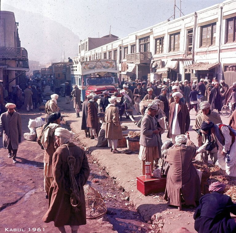A bustling street scene in Kabul from 1961, with people in traditional Afghan clothing engaging in daily activities among vehicles and street vendors.