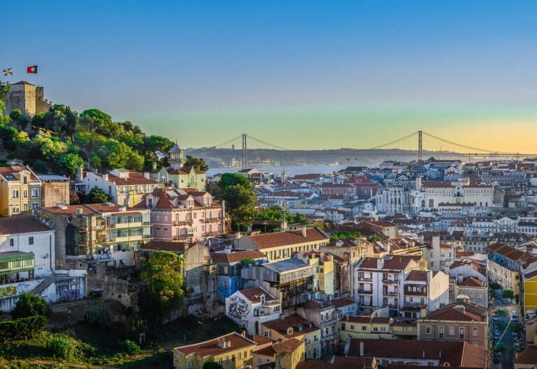 A panoramic view of Lisbon at sunset with the castle on the left, the 25 de Abril Bridge in the background, and the city's traditional architecture in the foreground.