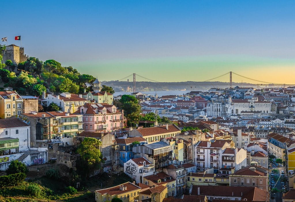 A panoramic view of Lisbon at sunset with the castle on the left, the 25 de Abril Bridge in the background, and the city's traditional architecture in the foreground.
