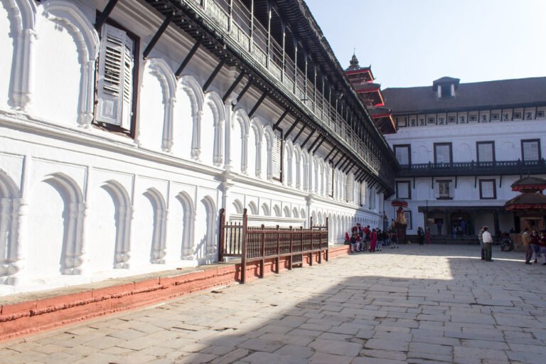 A sunny view of an expansive courtyard with traditional architecture, featuring a white building with a series of arches and carved wooden balconies, and people gathered in the open space.