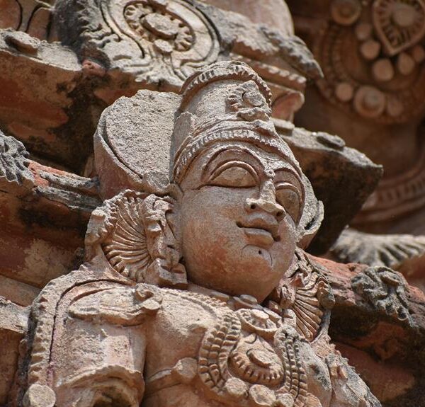 Stone sculpture of a deity's face with ornate headgear and jewelry, set against a temple wall with intricate carvings.