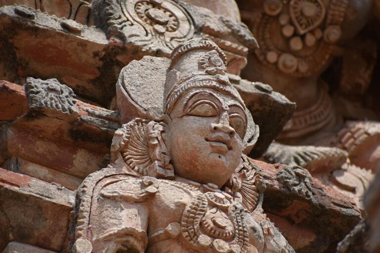 Stone sculpture of a deity's face with ornate headgear and jewelry, set against a temple wall with intricate carvings.