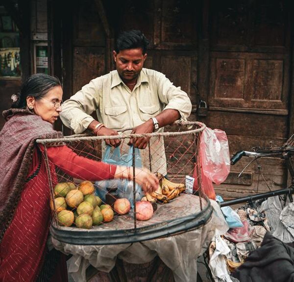 A woman in a red saree is purchasing fruit from a street vendor who has his goods displayed on a cart attached to a bicycle.