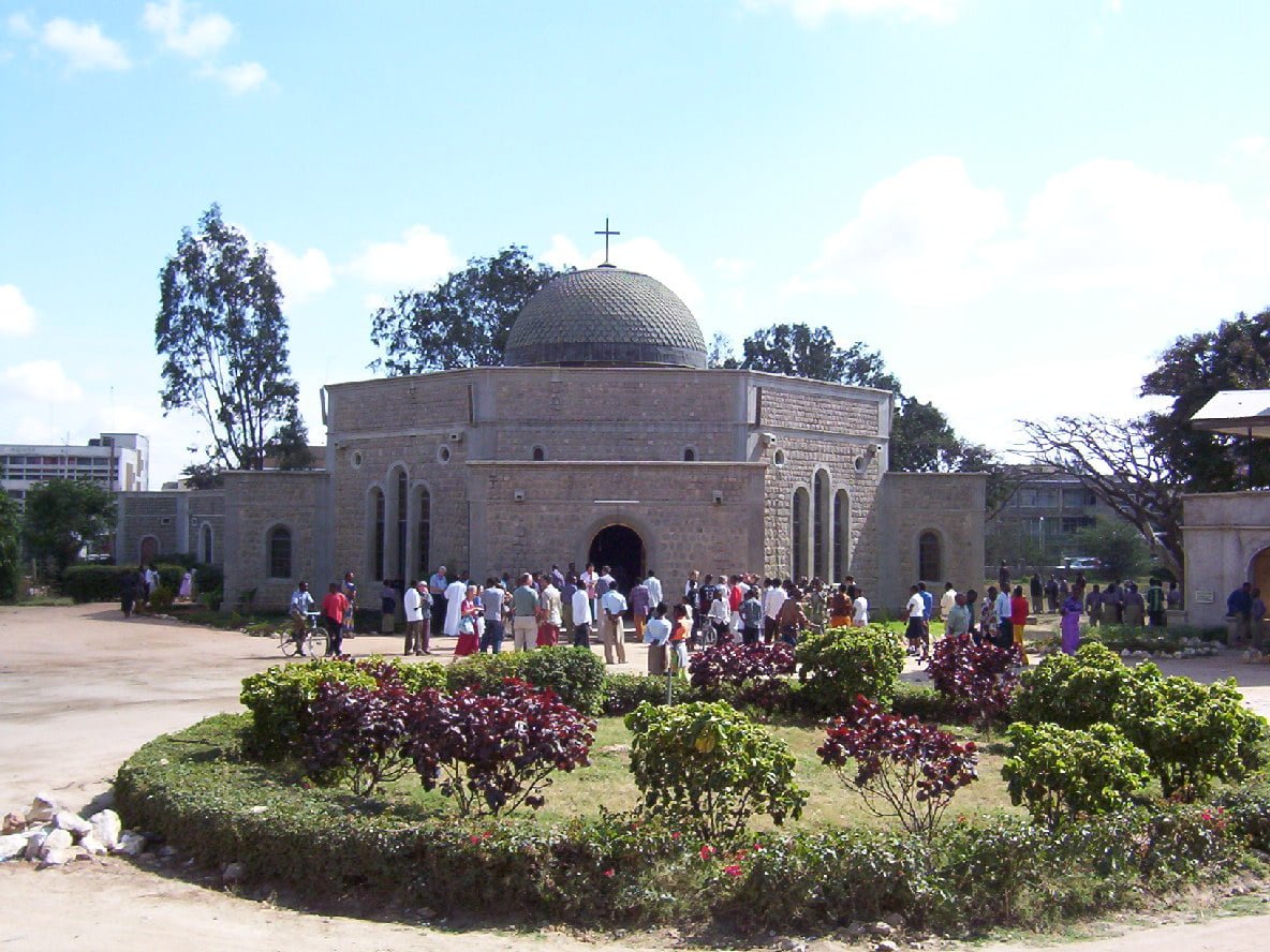 A stone church with a dome and cross, surrounded by people, under a clear sky with shrubbery in the foreground.