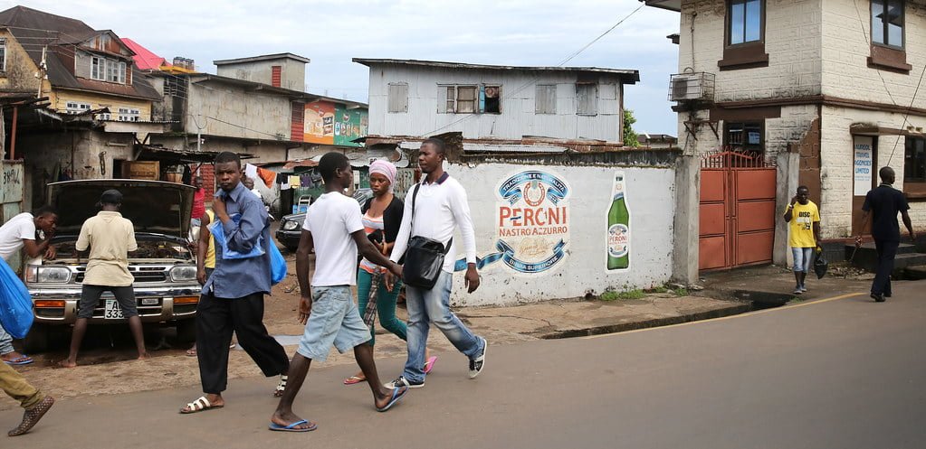 People walking along a busy street in an urban area with rundown buildings and a car being repaired, and advertisements on a perimeter wall.