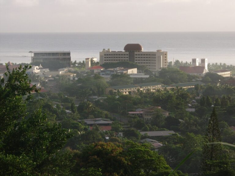A coastal cityscape with various buildings, including a distinctive building with a red dome, set against a backdrop of a calm sea and overcast sky.