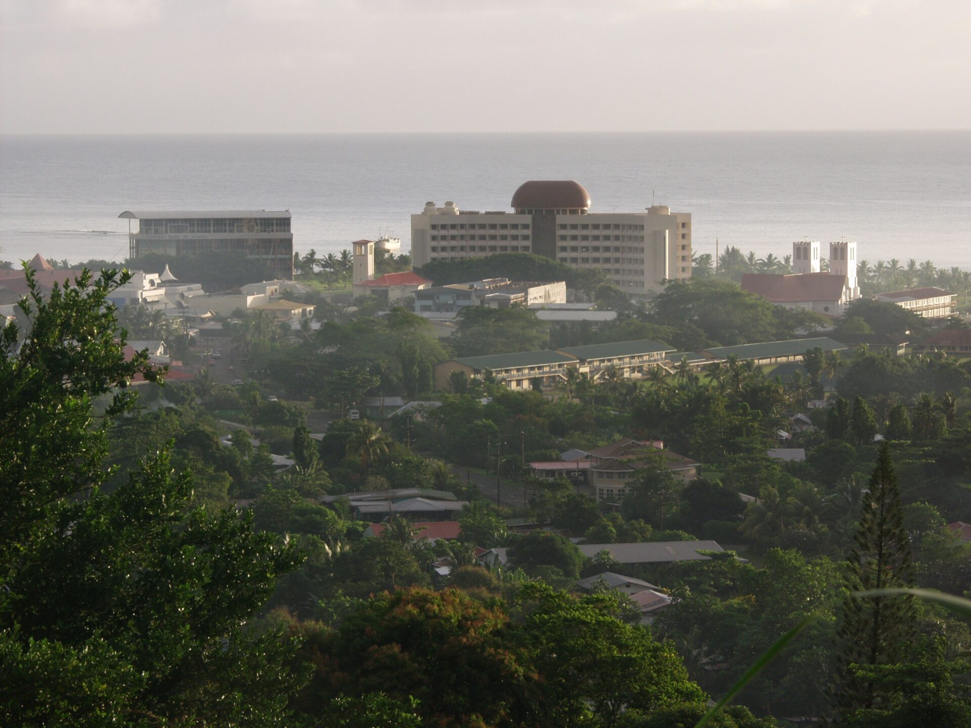 A coastal cityscape with various buildings, including a distinctive building with a red dome, set against a backdrop of a calm sea and overcast sky.