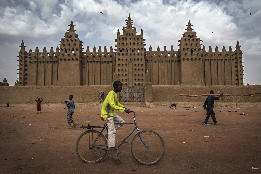 A man rides a bicycle in front of the towering walls of the Great Mosque of Djenné, Mali, with other individuals and a goat scattered in the foreground.