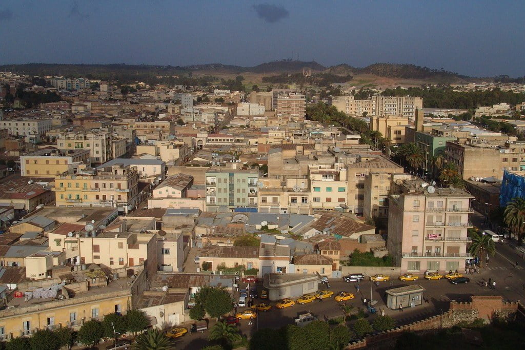 Aerial view of a dense urban area with a mix of mid-rise buildings, brightly colored taxis on the streets, and distant hills under a cloudy sky.