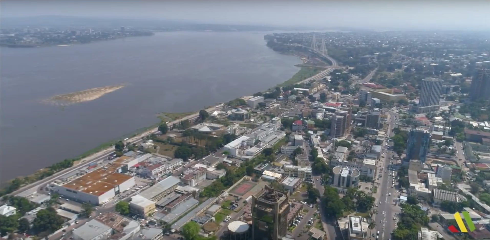 Aerial view of a cityscape next to a wide river, with a bridge in the distance and a sandbank visible in the water.