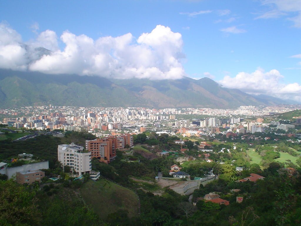 A panoramic view of a city nestled in a valley with tall buildings surrounded by green areas and mountains partially covered by clouds in the background.