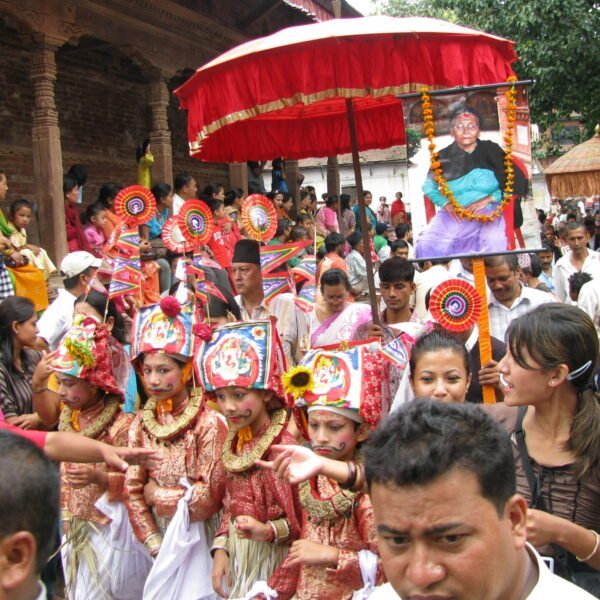 A vibrant procession with a group of young children in traditional Nepalese costumes and painted faces, carrying colorful pinwheels and cultural artifacts, followed by a large photo of a smiling individual adorned with flowers. A crowd of onlookers surrounds the procession in an outdoor setting with a historical building in the background.