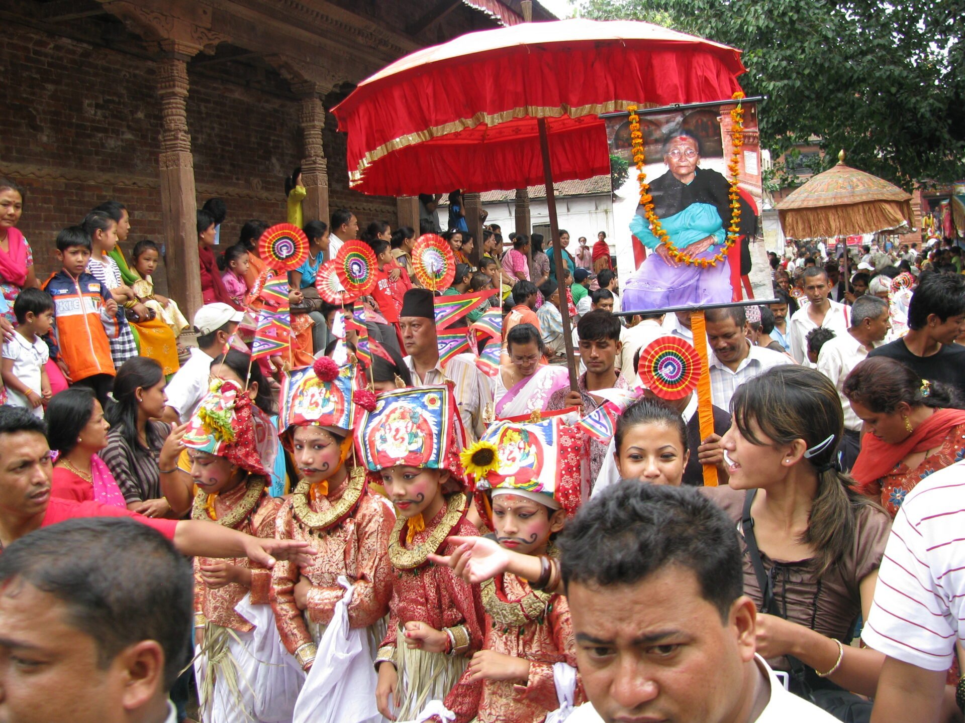 A vibrant procession with a group of young children in traditional Nepalese costumes and painted faces, carrying colorful pinwheels and cultural artifacts, followed by a large photo of a smiling individual adorned with flowers. A crowd of onlookers surrounds the procession in an outdoor setting with a historical building in the background.
