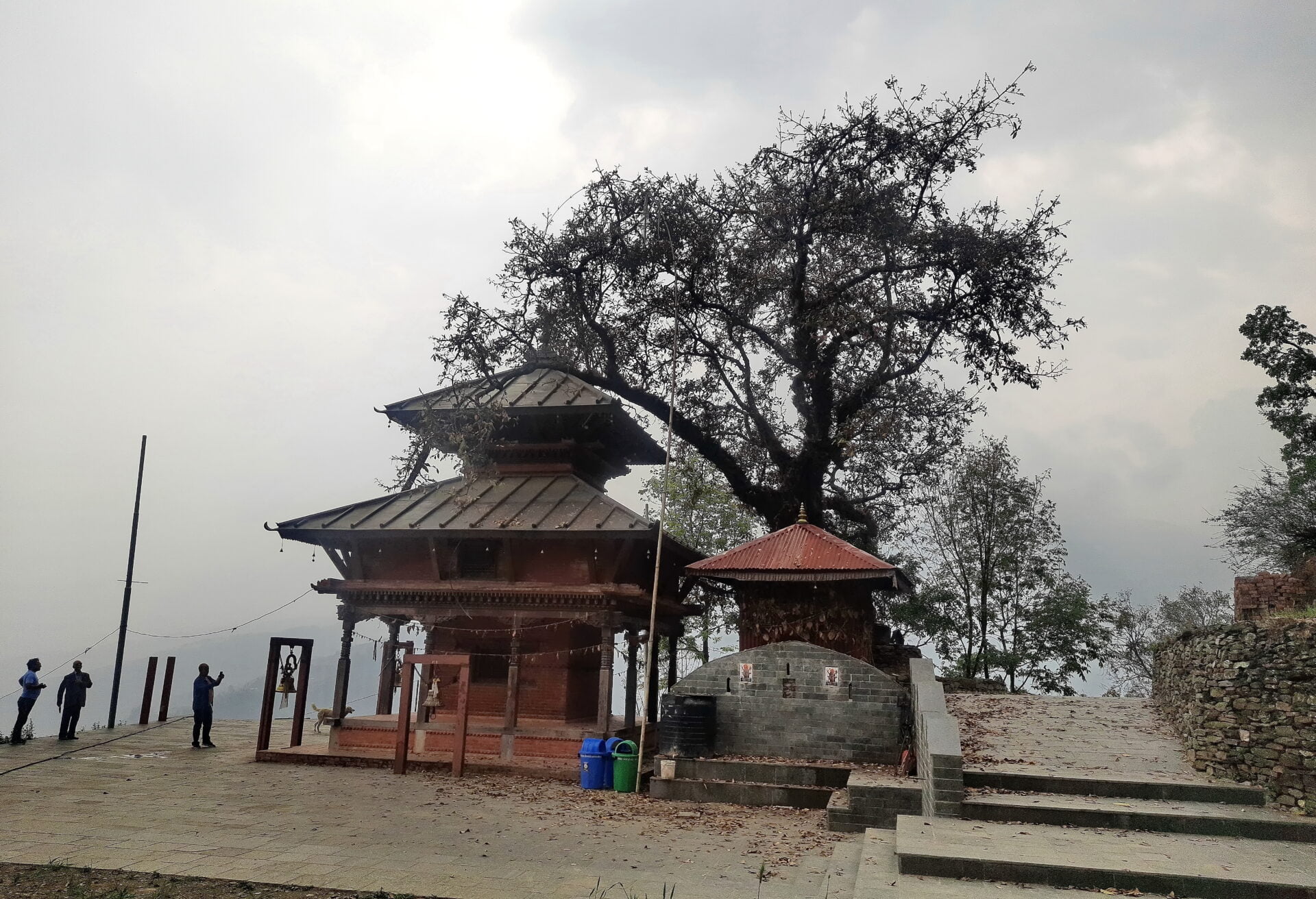 Alt text: A traditional temple with a pagoda-style roof surrounded by trees under a hazy sky. There are a few people nearby and stone steps leading up to the buildings.