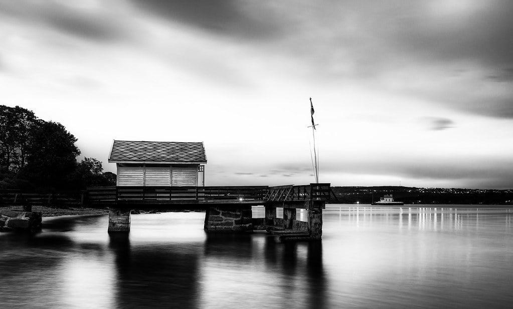 A black and white photograph of a small boathouse on a calm body of water with smooth long-exposure effects, under a cloudy sky.