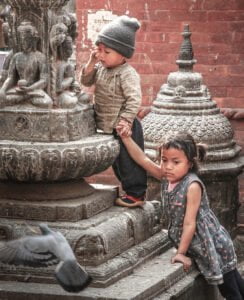 Two young children are playing near an intricately carved stone fountain featuring a Buddha statue, with a blurred pigeon in flight in the foreground. The boy is touching his face and the girl is looking away, both are surrounded by traditional Nepalese architecture.
