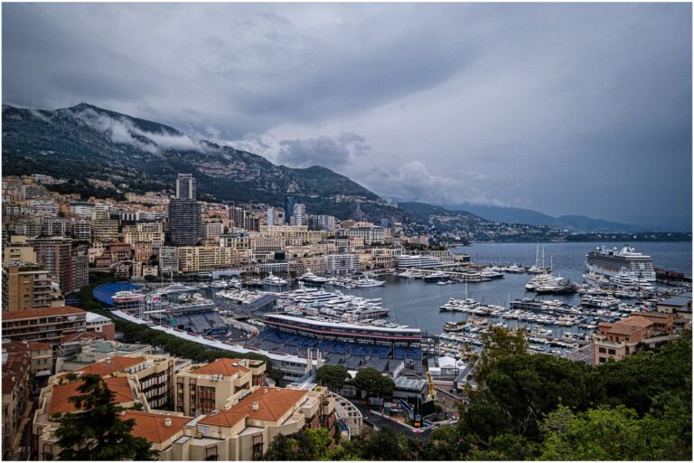 An overcast view of Monaco's marina with luxury yachts, surrounded by densely packed urban buildings, backed by mountains partially hidden by low clouds.