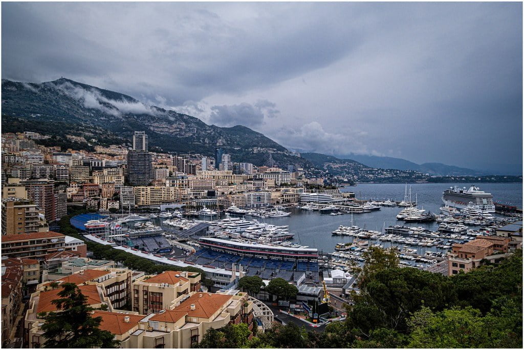 An overcast view of Monaco's marina with luxury yachts, surrounded by densely packed urban buildings, backed by mountains partially hidden by low clouds.