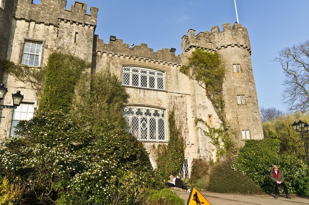 A medieval castle with ivy-covered walls, ornate windows, and a tower under a blue sky; flowering shrubs in the foreground with people nearby, one sitting and one standing, next to a caution sign on the path.