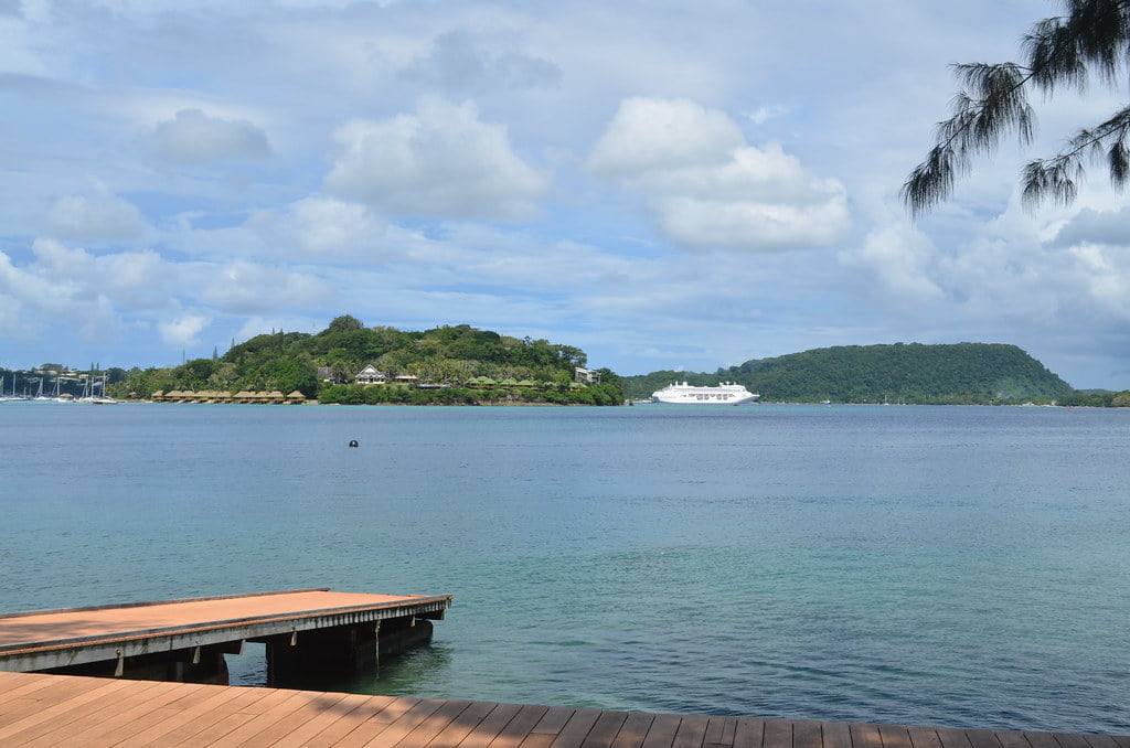 A tranquil seascape with a wooden pier in the foreground, a white yacht anchored near a lush, green island under a partly cloudy blue sky, and tree branches gently arching into the frame from the top right corner.
