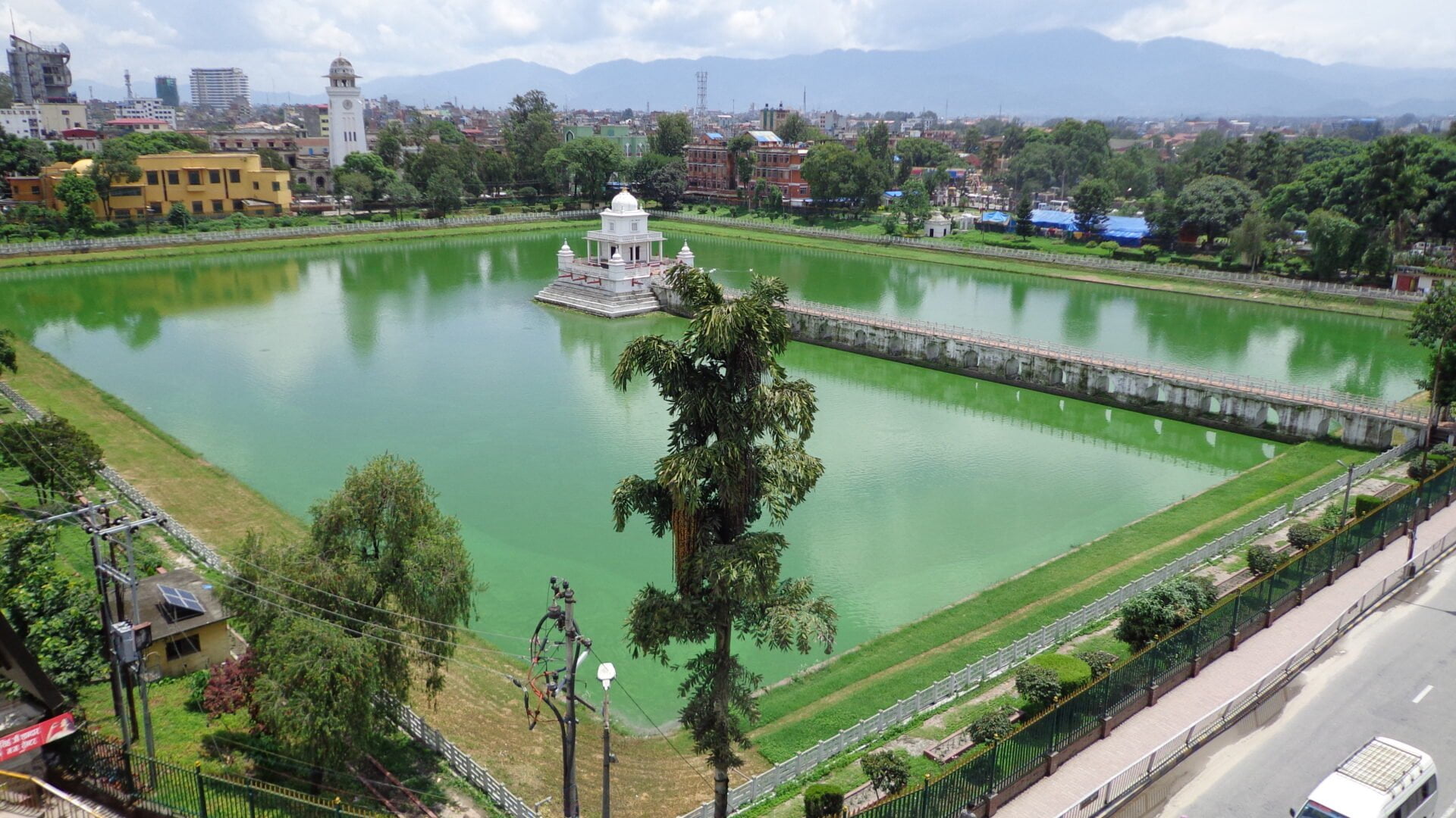 Aerial view of Rani Pokhari, an historic artificial pond, with a temple in the center, surrounded by green water and nestled in an urban landscape with streets, trees, and buildings in the background, under a cloudy sky.