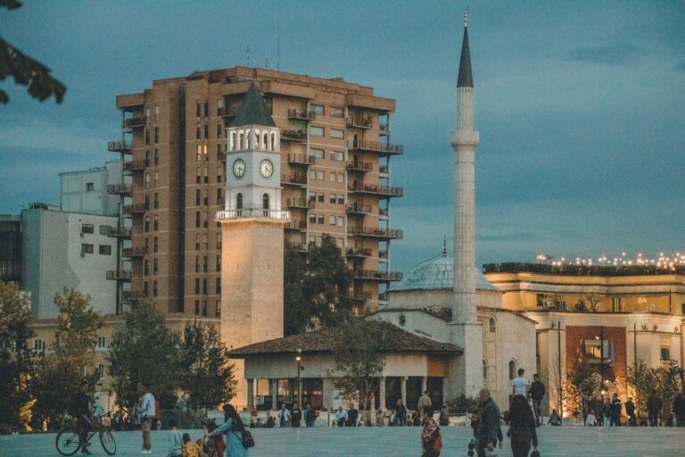 An evening view of an urban square with a clock tower, mosque minaret, and large residential building, with people walking and socializing.