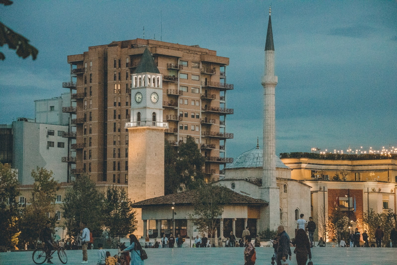 An evening view of an urban square with a clock tower, mosque minaret, and large residential building, with people walking and socializing.