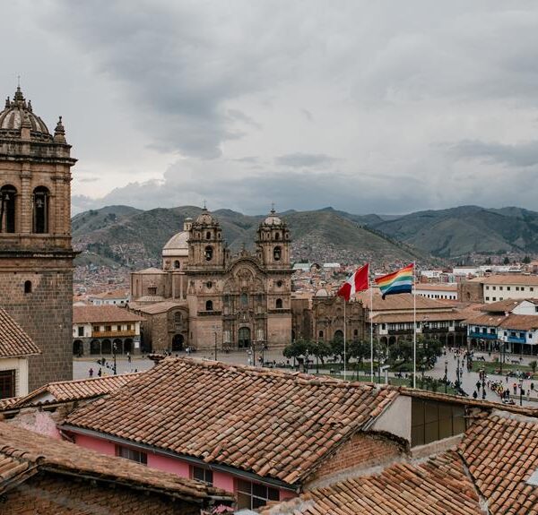 Overcast view of a historic city square with colonial architecture, distinctive church façades, and a Peruvian flag, with mountains in the background and red-tiled rooftops in the foreground.