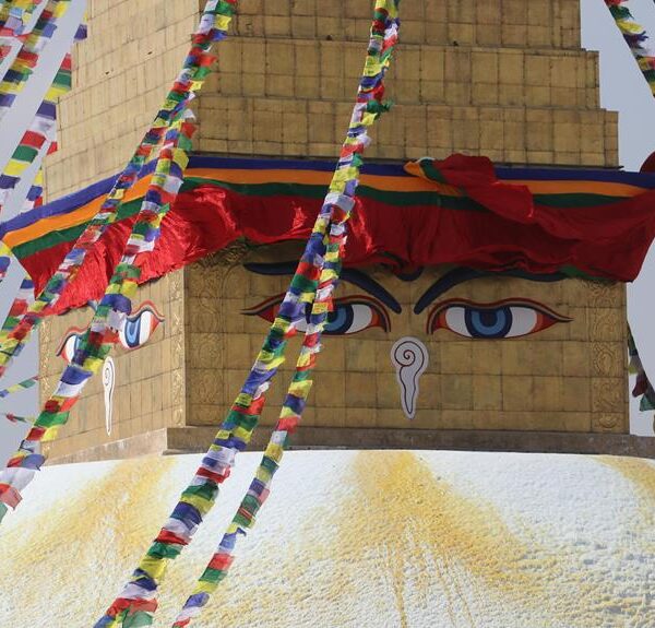 Prayer flags radiate from the golden apex of the Boudhanath Stupa in Kathmandu, Nepal, with the iconic painted eyes gazing outward beneath a vibrant red and blue canopy.