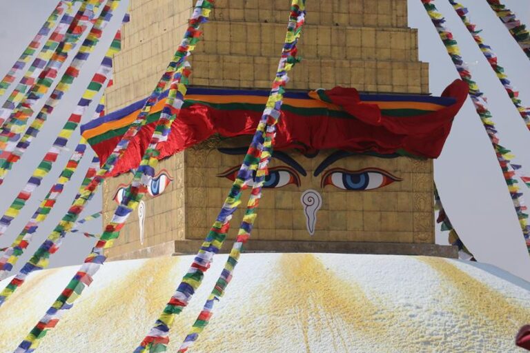 Prayer flags radiate from the golden apex of the Boudhanath Stupa in Kathmandu, Nepal, with the iconic painted eyes gazing outward beneath a vibrant red and blue canopy.