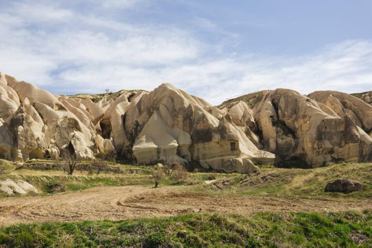 Eroded rock formations with various hues and patterns under a blue sky with scattered clouds in a grassy landscape.
