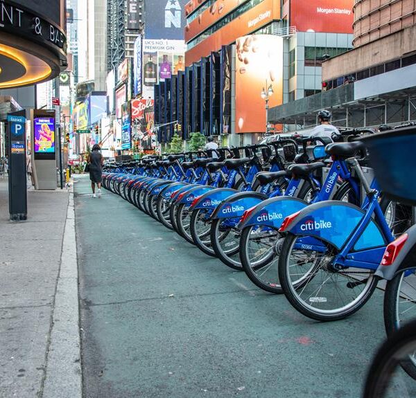 A row of blue Citi Bikes lined up at a docking station on a bustling city street with illuminated billboards and pedestrians in the background.