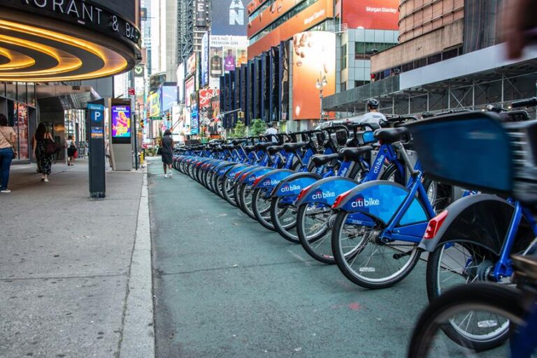 A row of blue Citi Bikes lined up at a docking station on a bustling city street with illuminated billboards and pedestrians in the background.