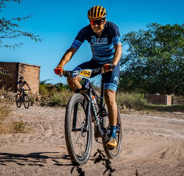 A cyclist in a blue and black racing kit leading a mountain bike race on a dirt path, with another competitor in the background, under a clear blue sky.