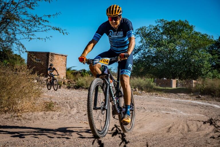 A cyclist in a blue and black racing kit leading a mountain bike race on a dirt path, with another competitor in the background, under a clear blue sky.