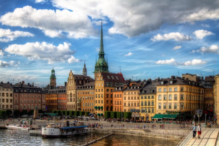 A scenic view of a European city waterfront with colorful buildings under a blue sky with clouds, featuring a spire, boats, and people walking along the quay.
