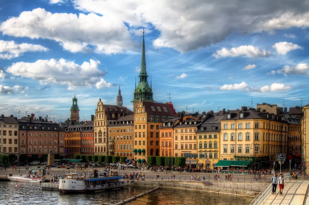 A scenic view of a European city waterfront with colorful buildings under a blue sky with clouds, featuring a spire, boats, and people walking along the quay.