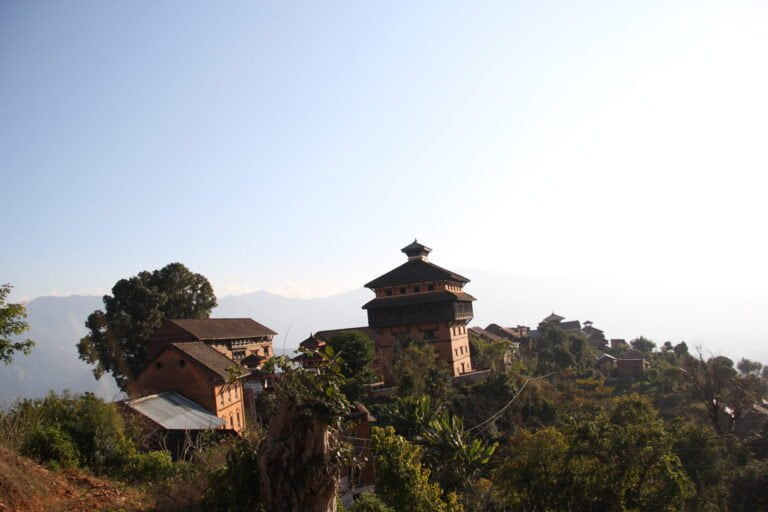 An ancient hilltop village with traditional multi-story buildings overlooking a mountain range under a clear sky.