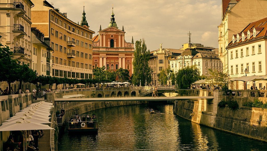 A scenic view of a European cityscape featuring a river with a boat, riverside terraces with umbrellas, elegant buildings, and a prominent pink church in the background, under a soft, golden twilight sky.