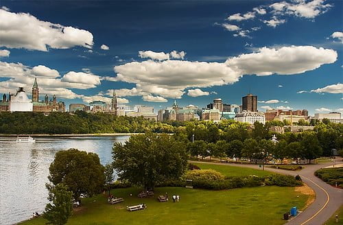 Skyline of a city with modern buildings beside a river, topped by a dramatic cloud-filled sky, viewed from a lush green park.